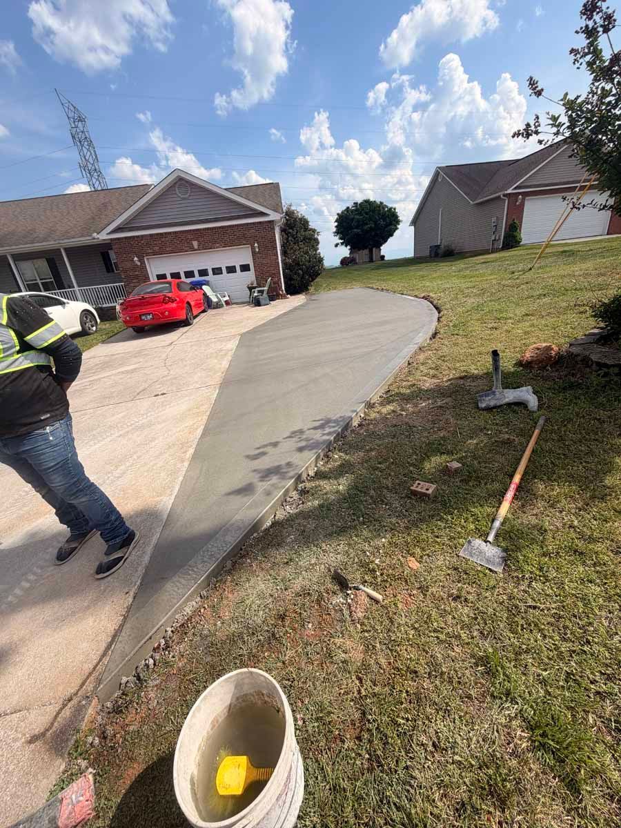 Suburban driveway under construction with a worker, red car, concrete path, and tools in the yard