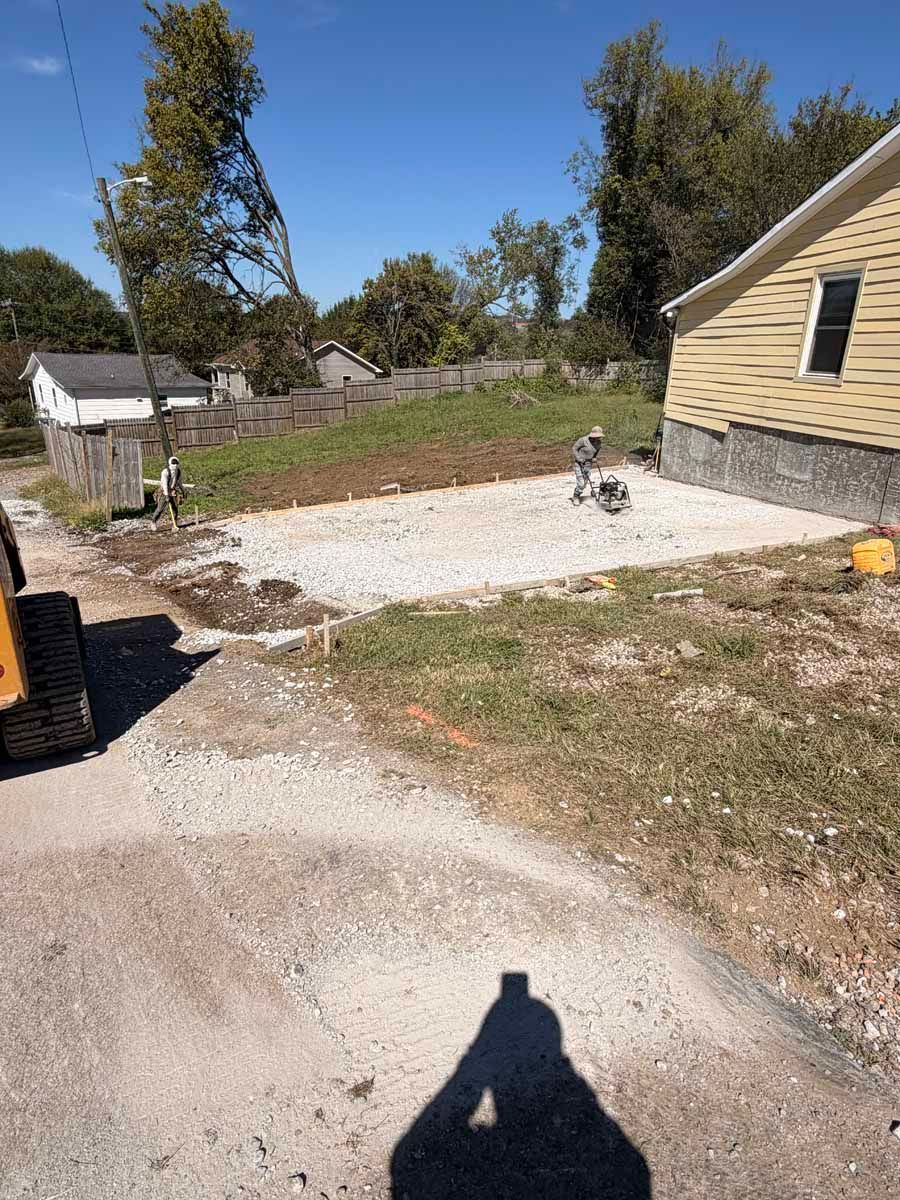 Rural yard with gravel driveway, small yellow house, trees, and a person riding a dirt bike near the house