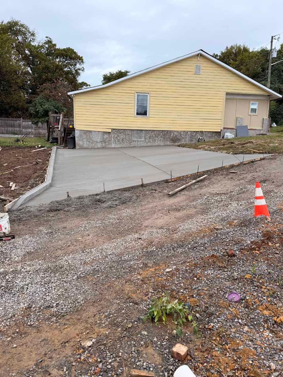 Yellow house with a new concrete driveway and gravel foreground, marked by an orange traffic cone.