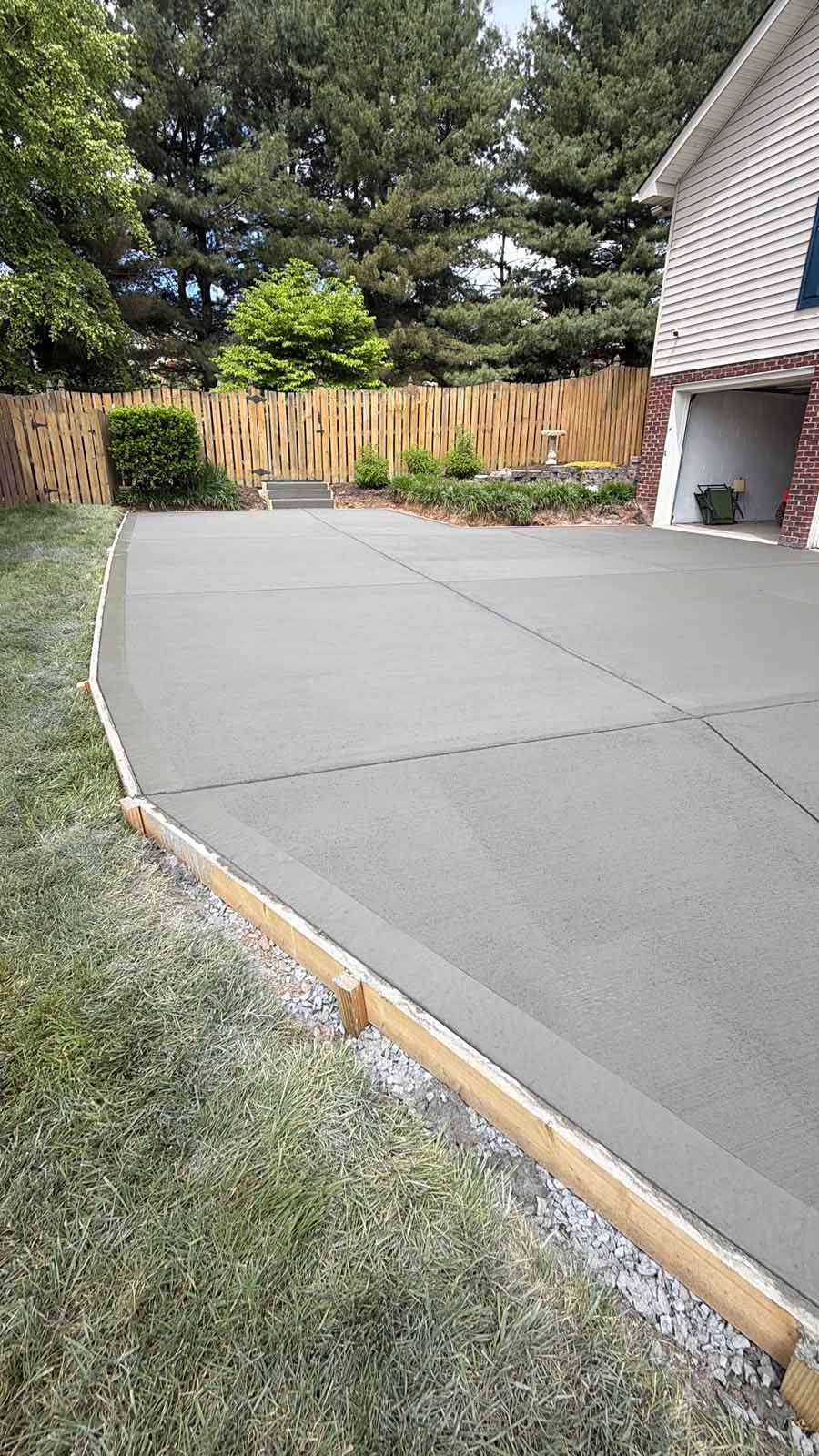 Freshly poured concrete patio beside a house, bordered by grass and a wooden fence, with trees in the background
