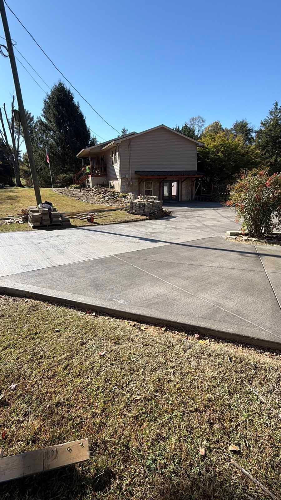 Driveway leading to a small house with a carport, bordered by grass and trees on a sunny day