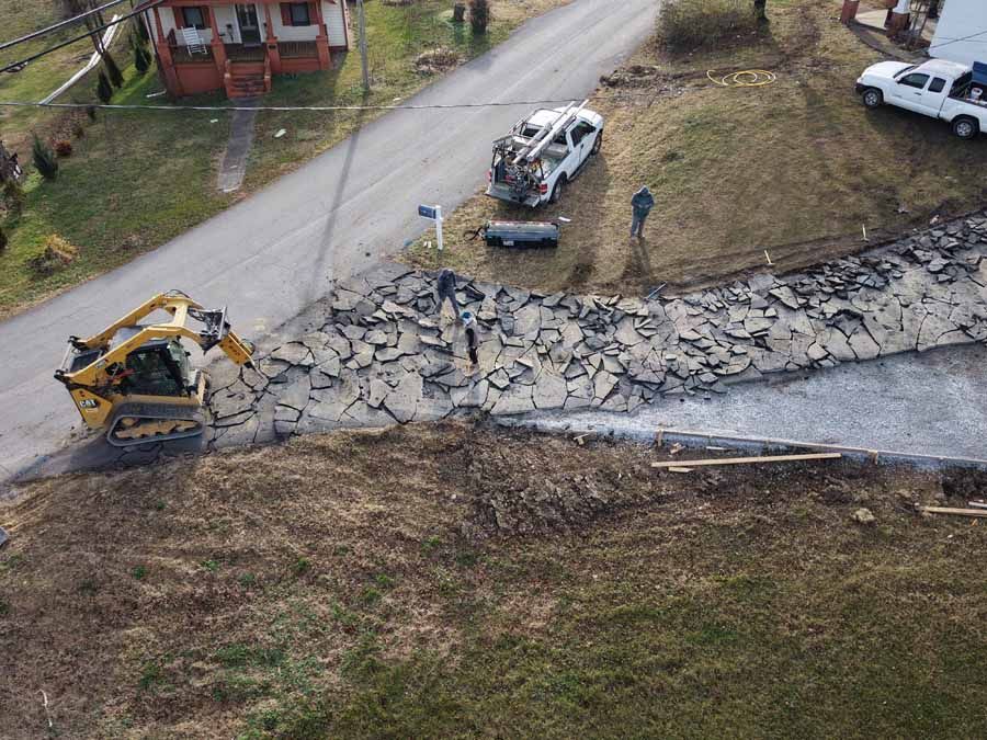 Workers repairing a collapsed roadside retaining wall beside a damaged road, with a backhoe and utility truck.