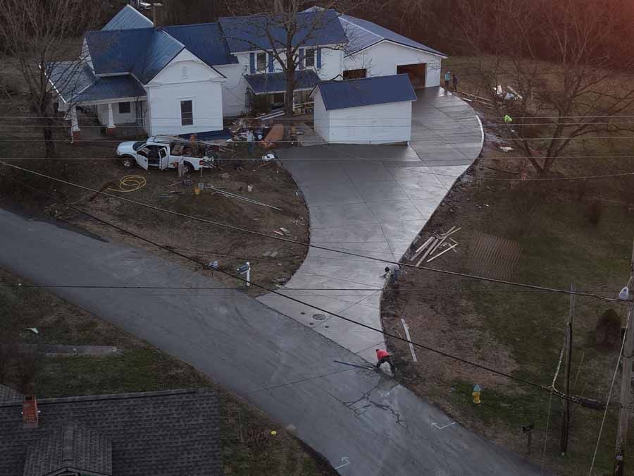 Aerial view of a house with a long driveway, parked vehicles, and a person in a red jacket near the road.