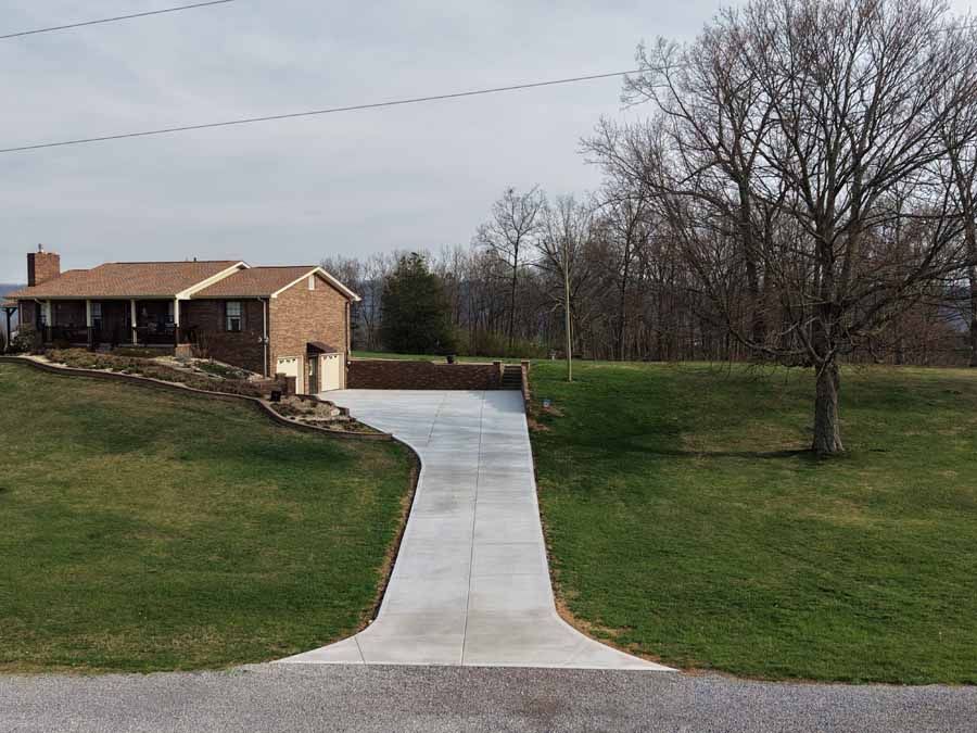Concrete driveway leading to a brick house with a porch, beside grassy slopes and leafless trees