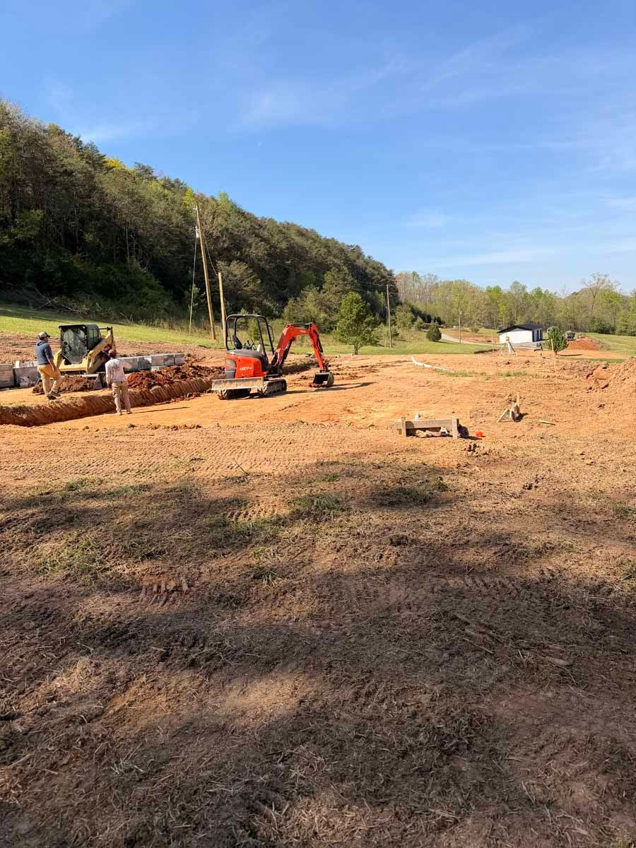Cleared dirt field with excavators and construction equipment beside a wooded hillside under a blue sky
