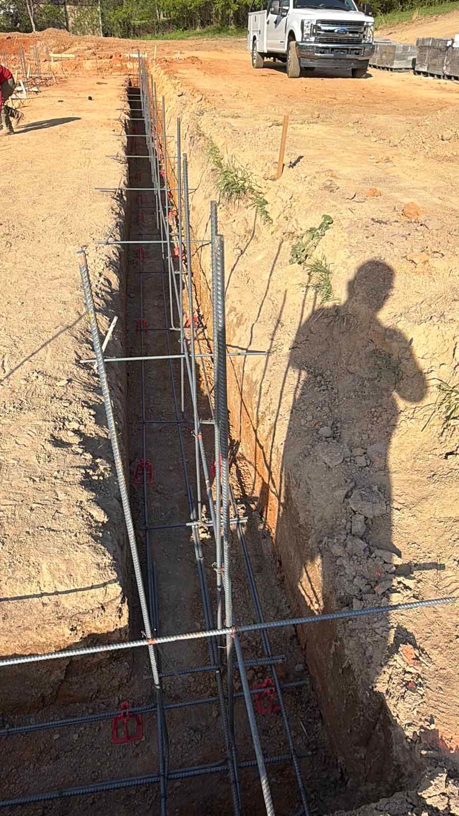 Construction trench with rebar and formwork on a dirt site, with a truck in the background.