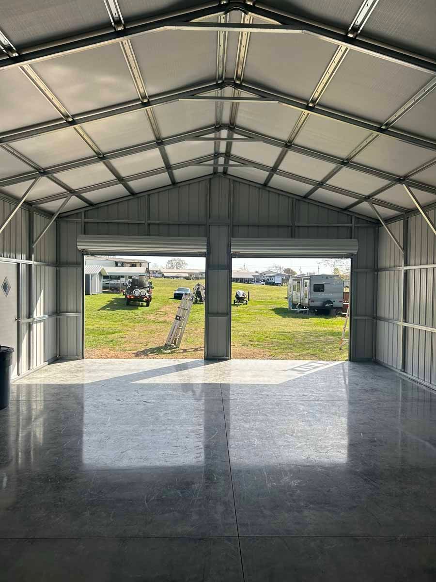 Empty metal garage with open front doors, showing a grassy yard and vehicles outside.
