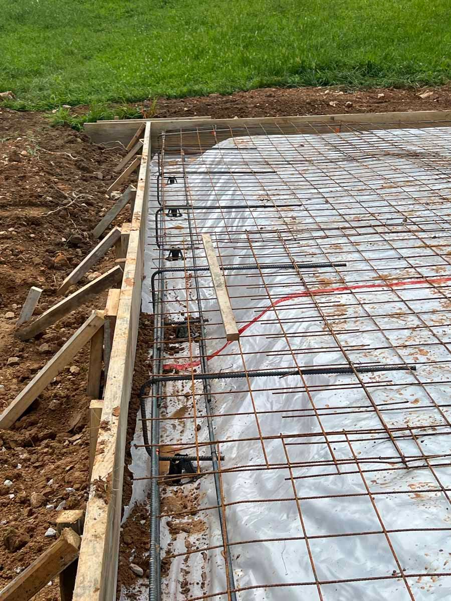 Construction site with steel rebar grid and plastic-covered foundation slab beside soil and grass