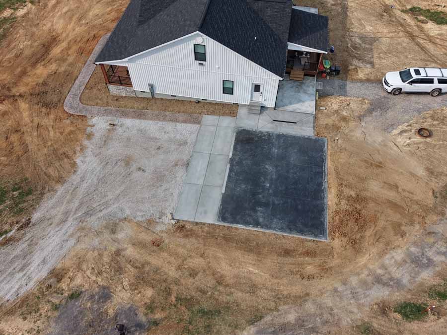 Aerial view of a white house with a black roof, driveway, and a parked white vehicle on a dirt lot.