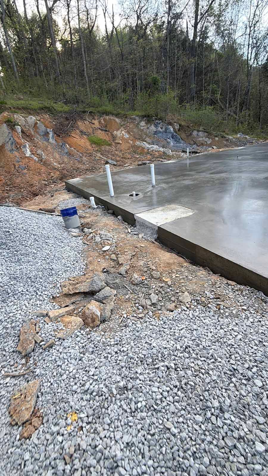 Fresh concrete slab with exposed rebar and gravel beside a wooded embankment