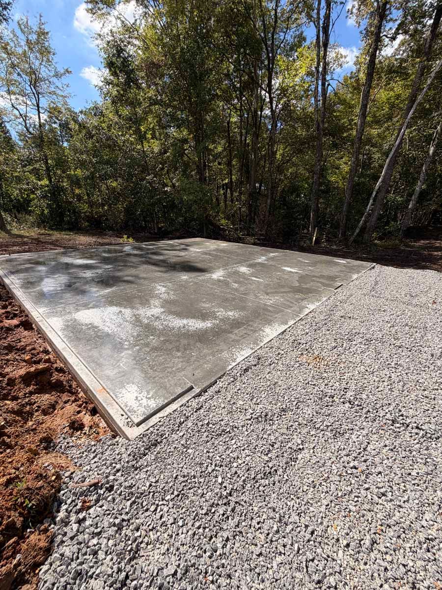 Fresh concrete slab beside a gravel driveway in a wooded outdoor area under sunlight