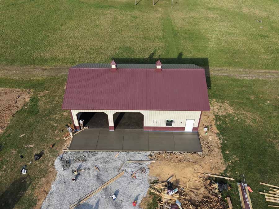 Aerial view of a small red-roofed building with a front porch beside a gravel area and grassy field.