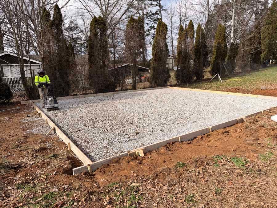 Gravel foundation pad under construction with a worker on the left in a wooded yard