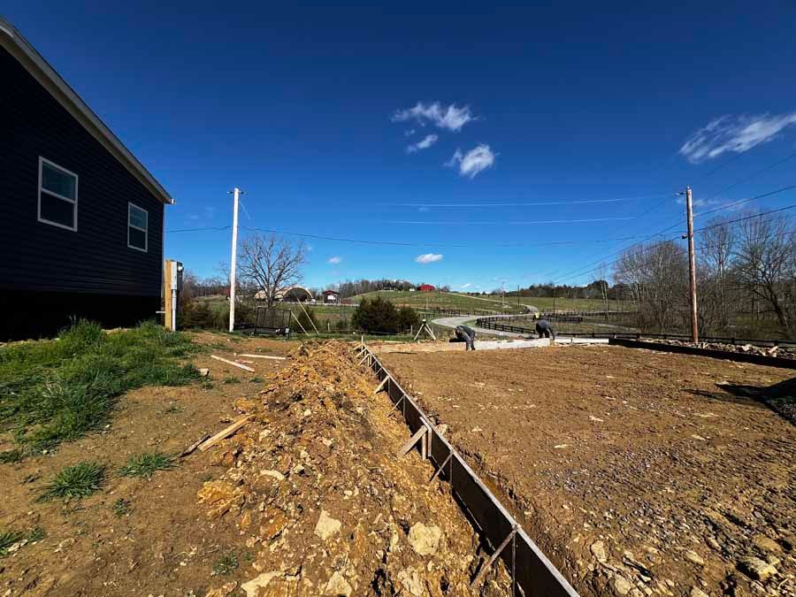 Graded construction site beside a dark house under a blue sky, with dirt and a concrete foundation outline