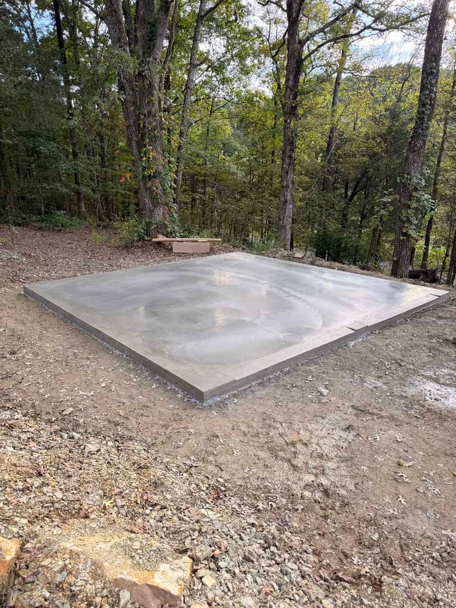 Fresh concrete slab set in a wooded clearing, with gravel and trees around it.