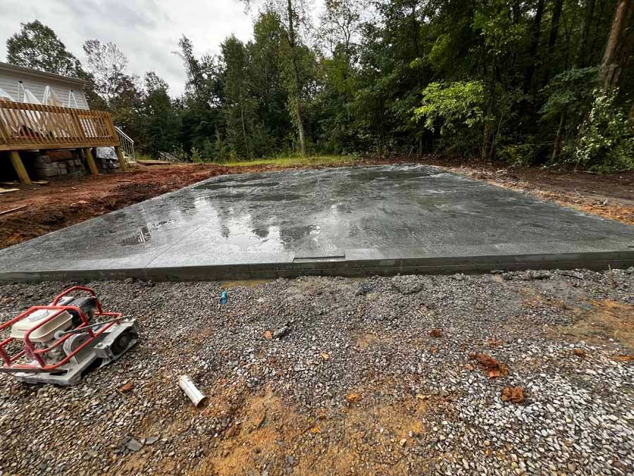 Fresh concrete slab on a gravel lot beside trees and a wooden structure under overcast sky