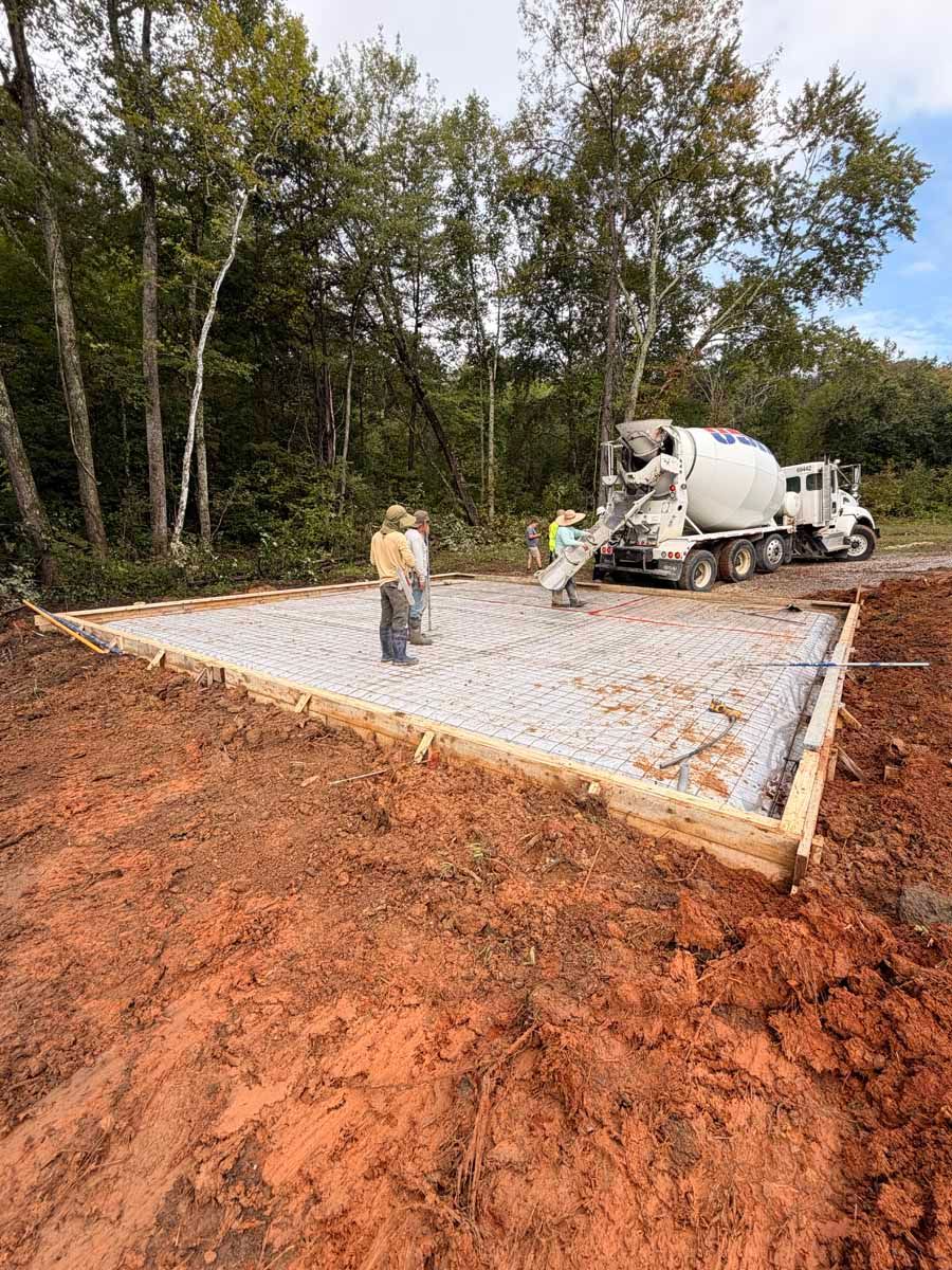 Concrete slab being poured in a wooded area, with a cement truck and workers on site.
