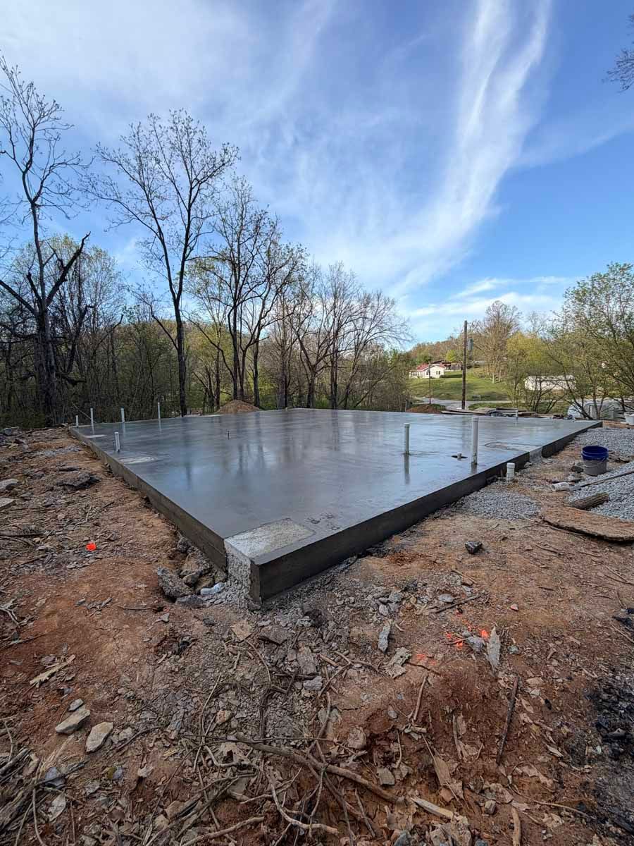 Fresh concrete slab foundation in a wooded clearing under a partly cloudy sky