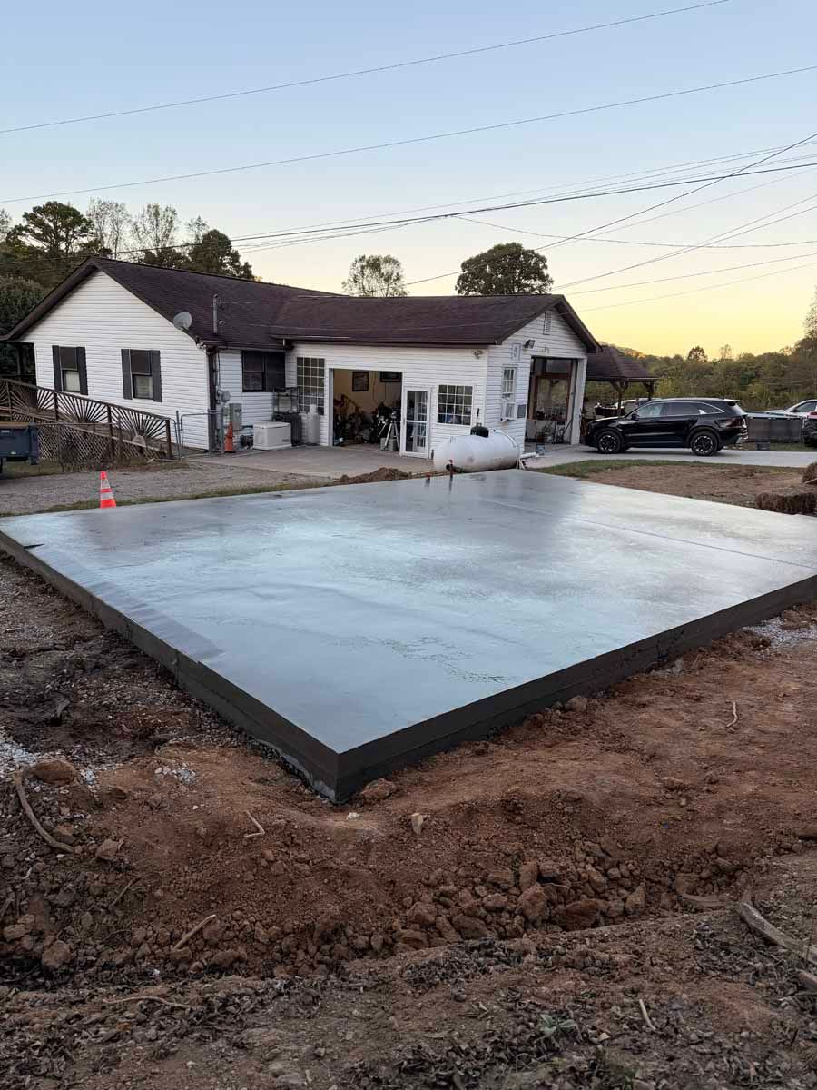 Concrete foundation slab in front of a white house at dusk, with dirt and construction materials nearby