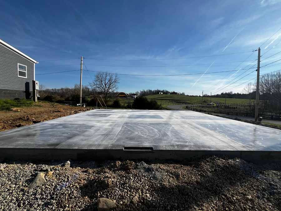 Concrete slab foundation on a gravel lot beside a house, under a clear blue sky.