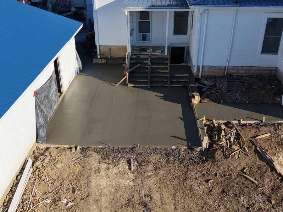 Fresh concrete slab beside a house, with steps leading to a white porch and dirt construction area nearby