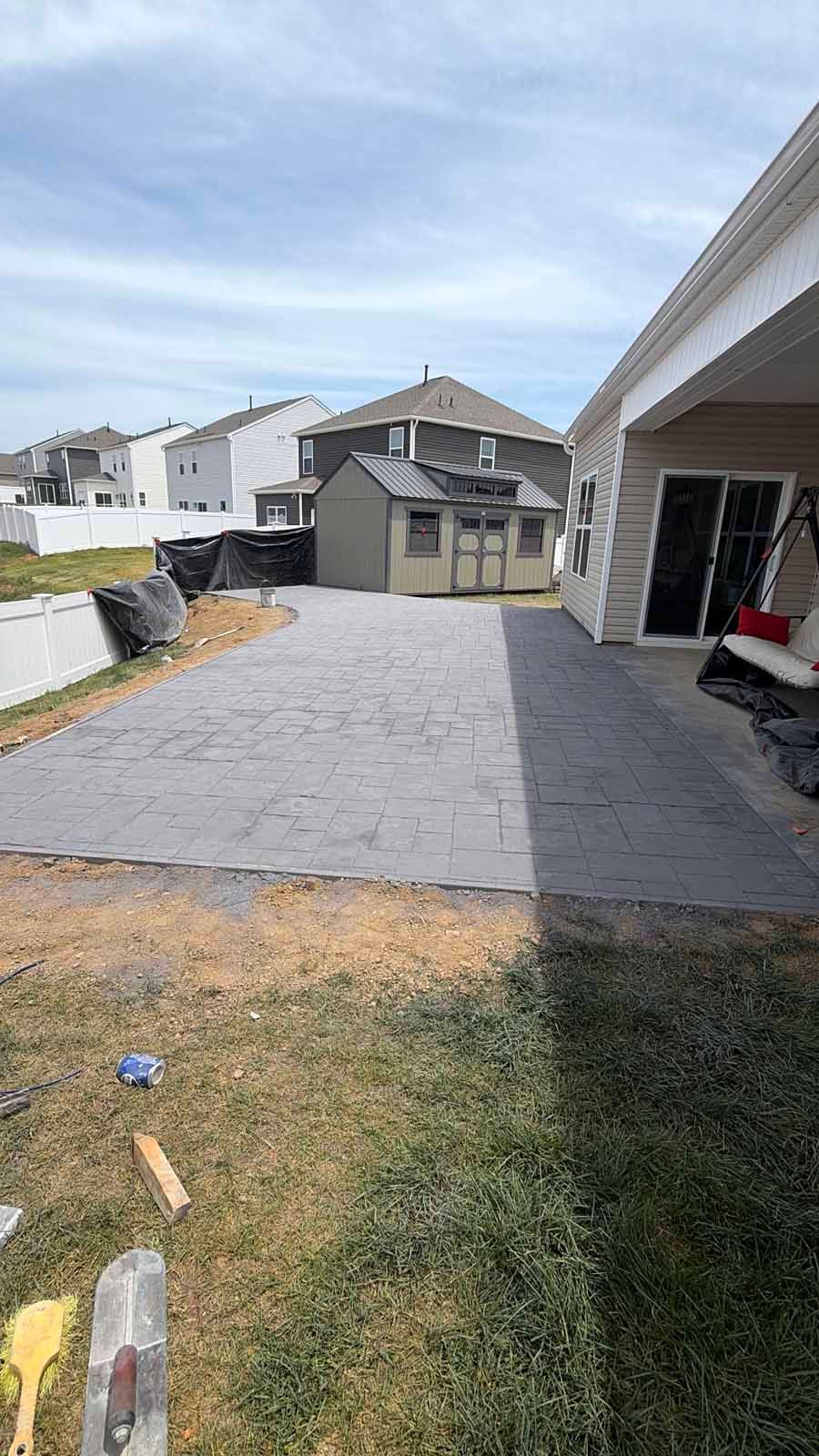 Backyard with a large gray paved patio beside a covered house patio and neighboring homes under a blue sky