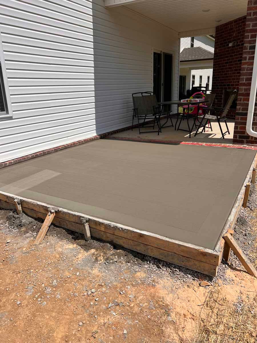 Fresh concrete patio slab with wooden forms beside a brick house and covered porch.