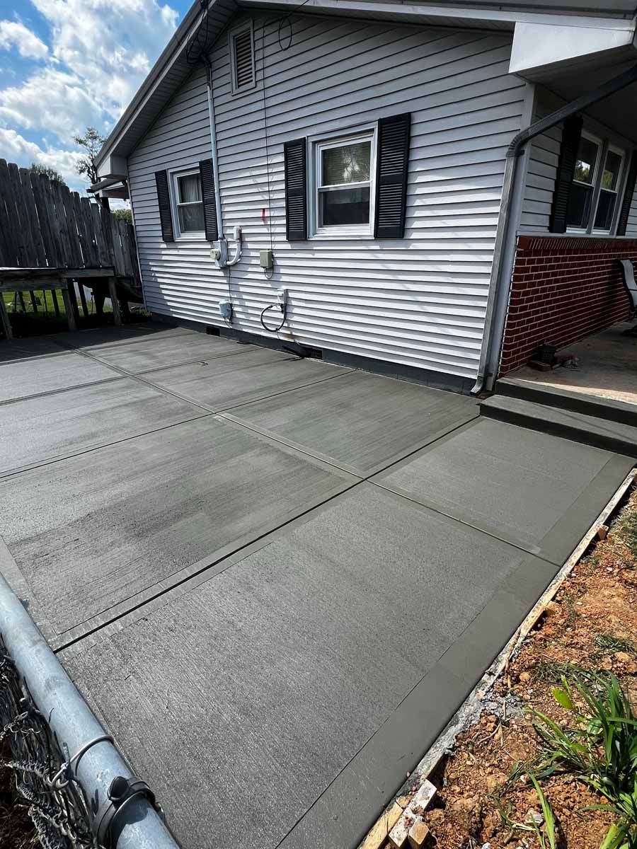 Concrete patio beside a gray house with steps and a railing, bordered by grass and fallen leaves