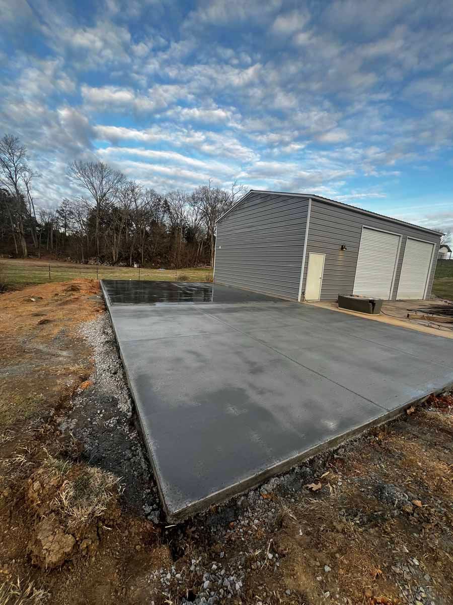 Concrete slab beside a gray shed under a cloudy sky, surrounded by bare trees and dirt ground