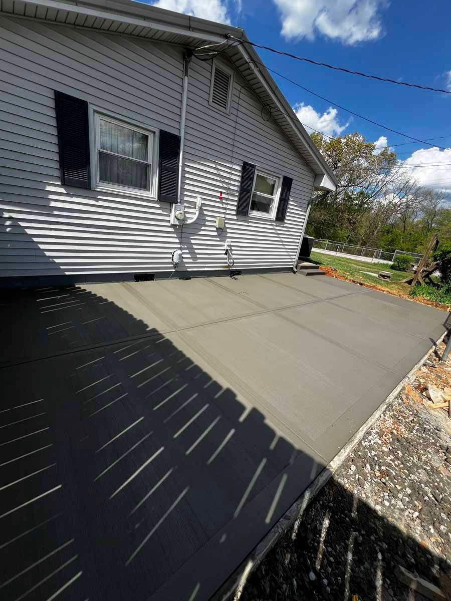 Backyard patio beside gray house with a large concrete slab and gravel edge under a blue sky.