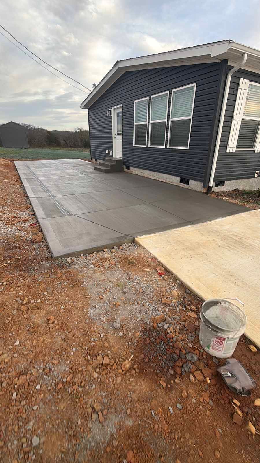Concrete patio beside a dark blue house with white trim, a gravel yard, and a paint bucket in the foreground