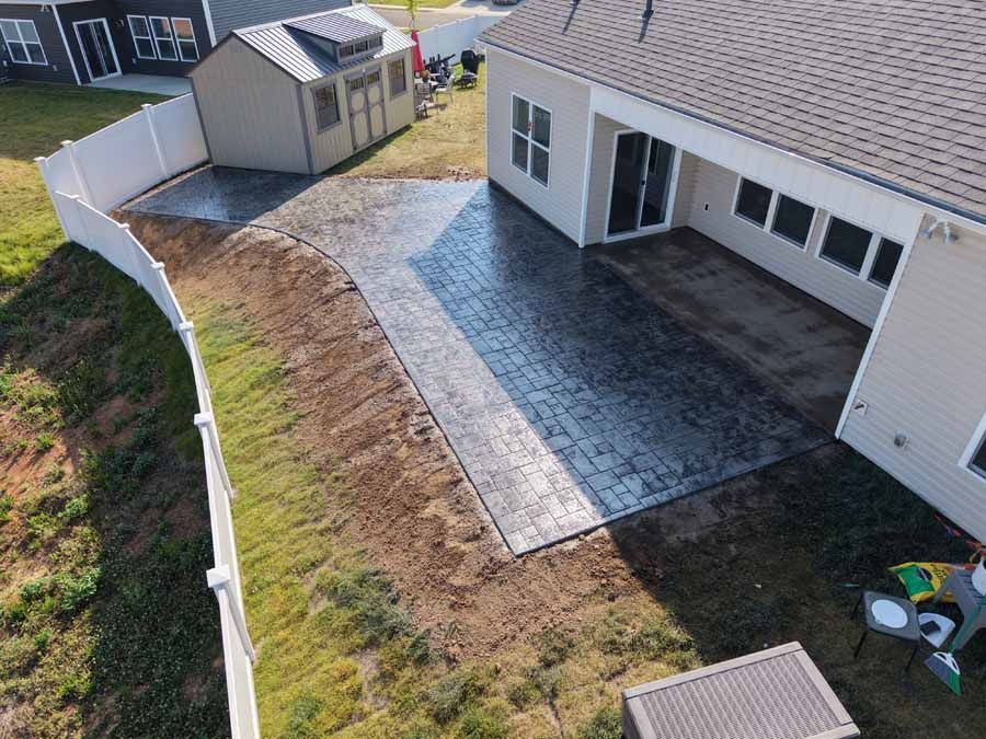 Backyard with white house, paved patio, shed, and curved white fence bordering a grassy lawn
