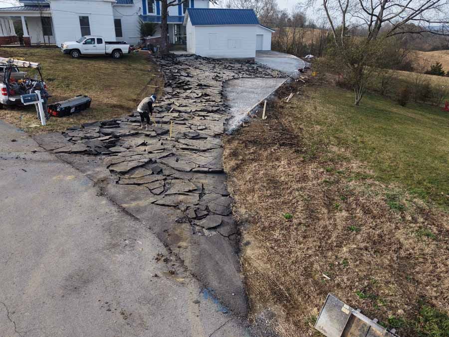 Damaged driveway and collapsed asphalt beside a house, with scattered debris and eroded ground after flooding