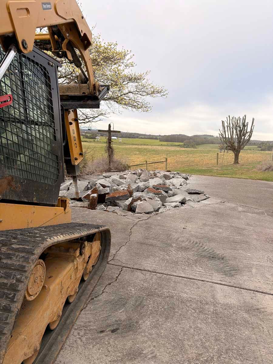 Bulldozer beside a pile of broken concrete on a rural roadside, with fields in the background