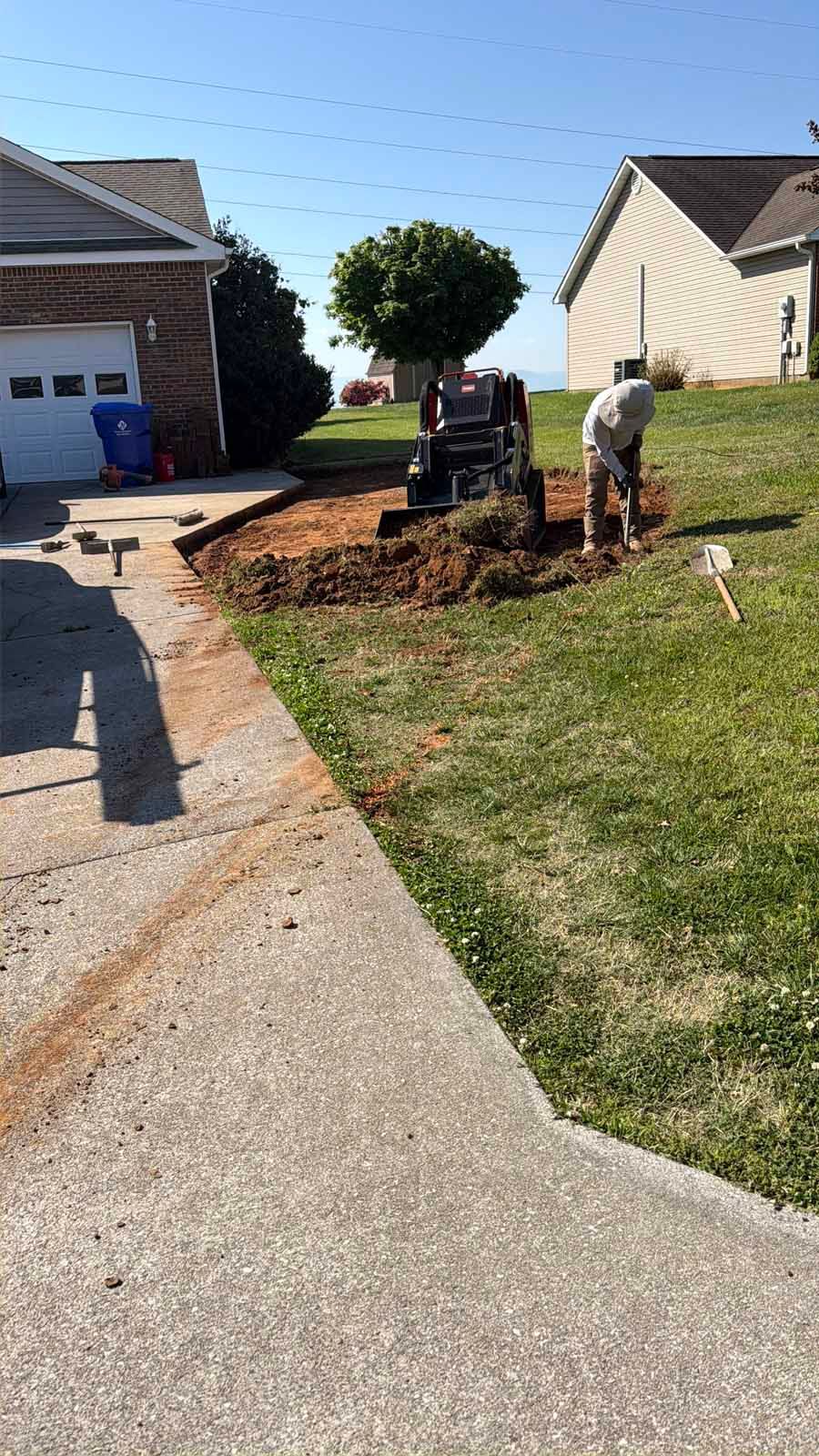 Two workers digging soil beside a driveway in a suburban front yard, with a small machine and piles of dirt.