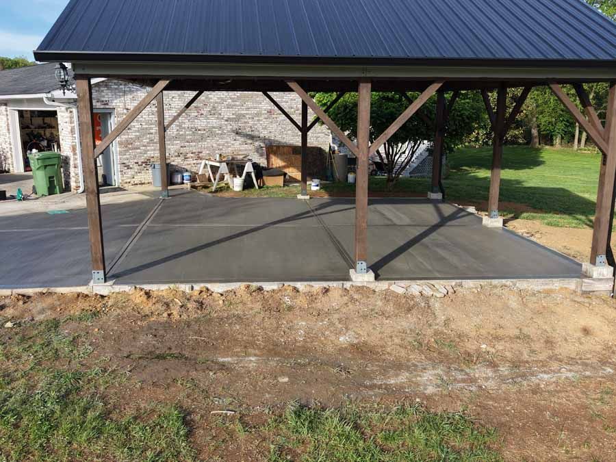 Covered concrete patio with metal roof and wooden posts beside a stone building.