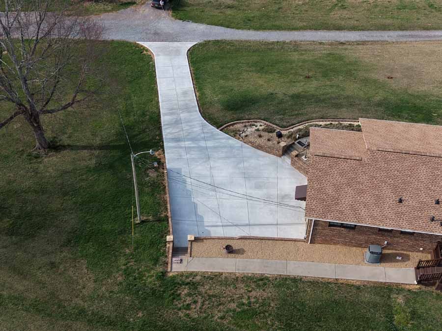Aerial view of a house with a long white driveway and brown roof beside a grassy yard.
