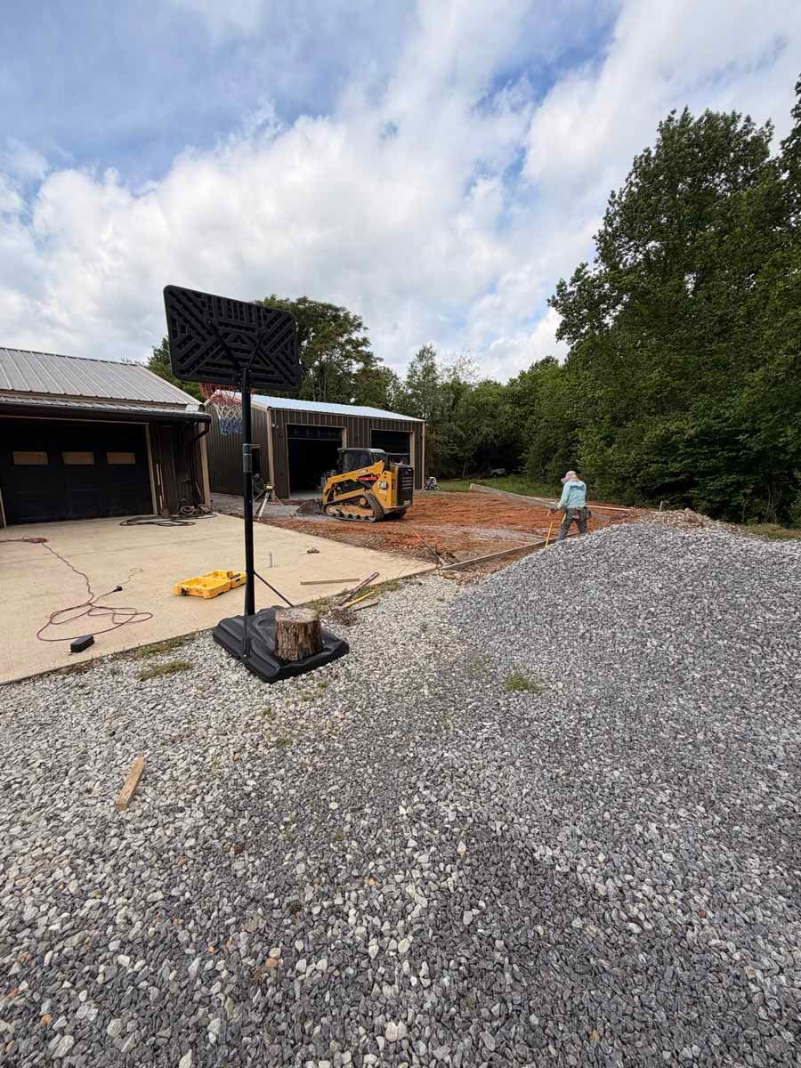 Construction site with gravel piles, a small excavator, and a person near a building under a cloudy sky