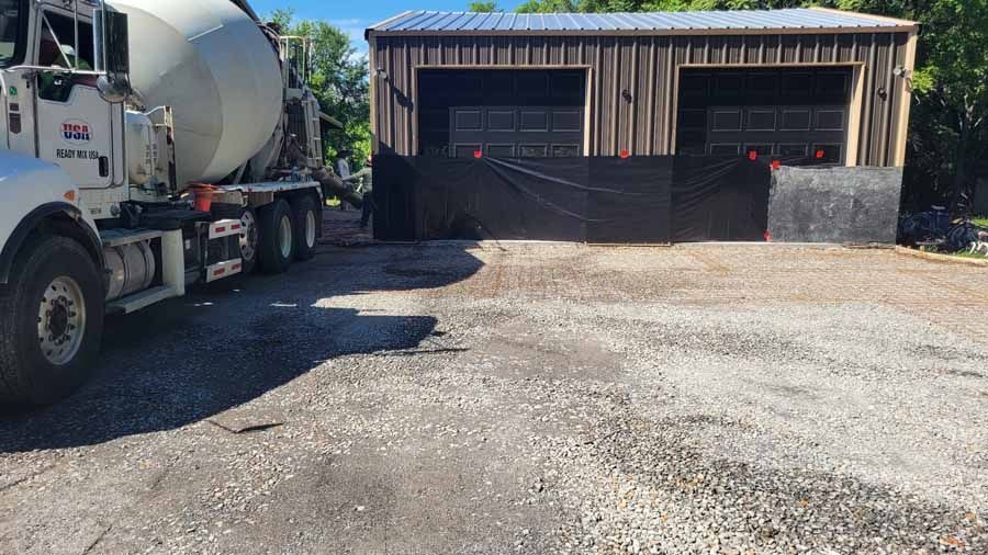 Concrete mixer truck beside a rustic barn with open bays and a gravel yard