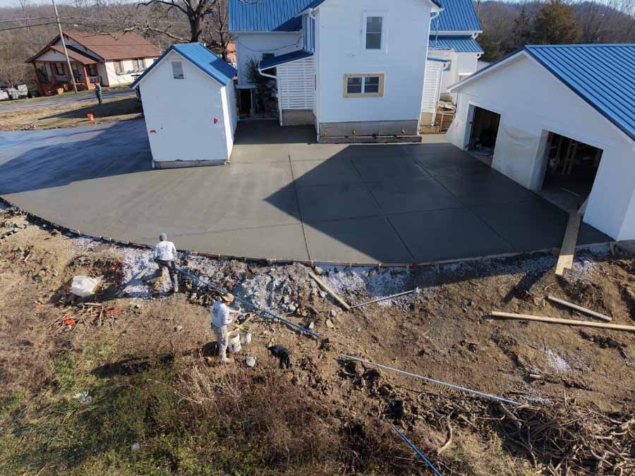 Aerial view of a house with a large concrete driveway and detached garage in a rural yard.