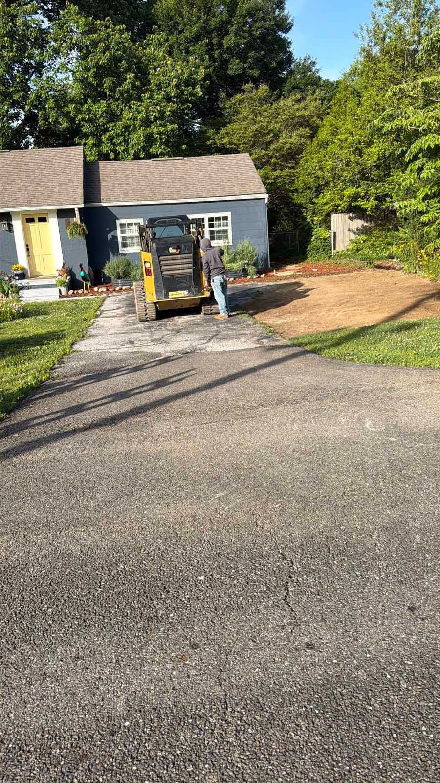 Gravel driveway with a small skid steer parked beside a white garage and trees in the background