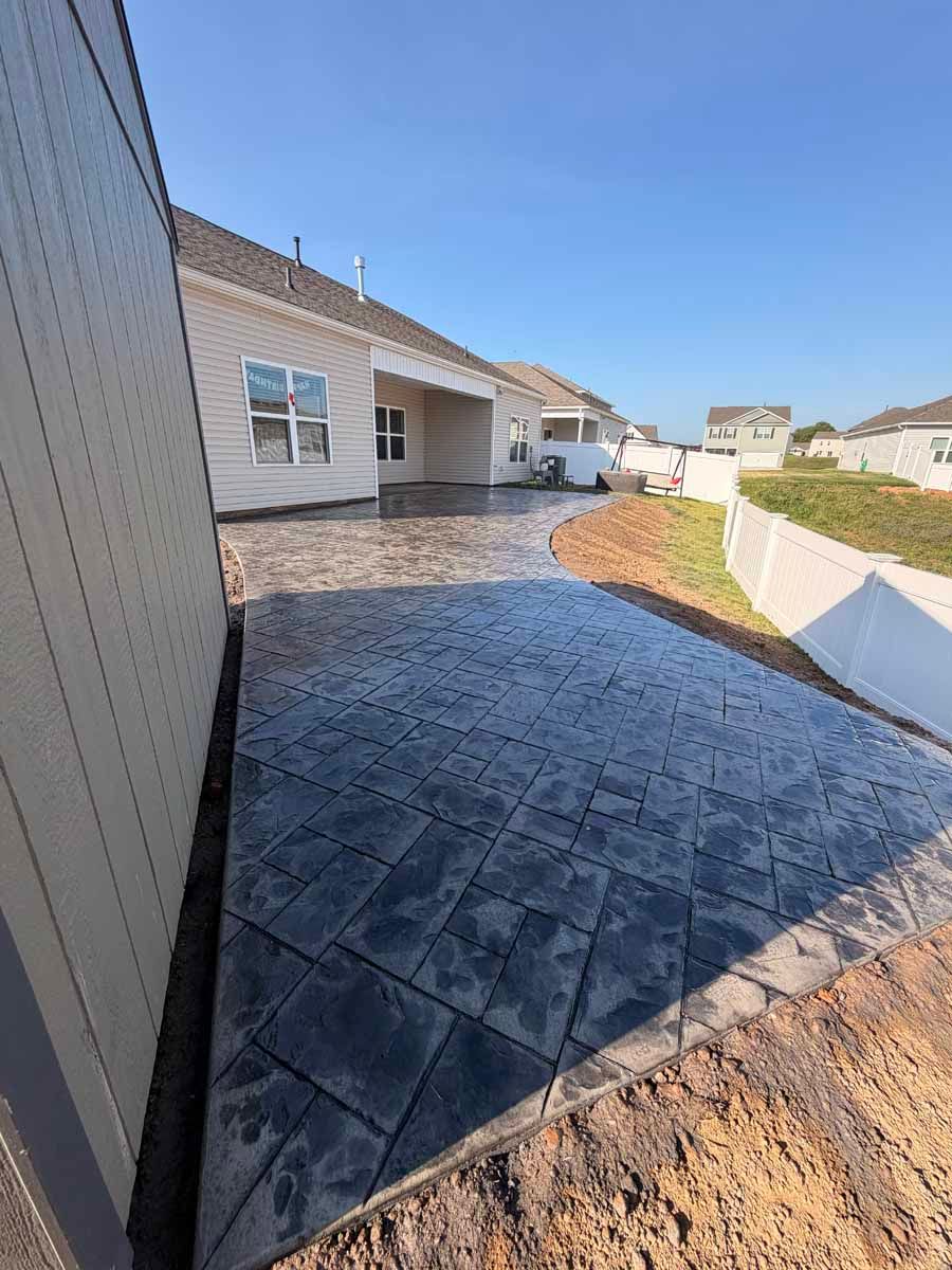 New home patio with dark pavers and beige siding, beside a gravel and landscaped yard under a blue sky