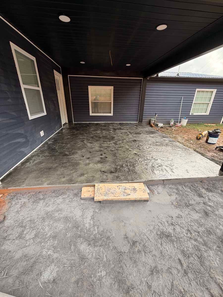 Covered backyard patio with dark siding, concrete floor, and two windows under a roof overhang