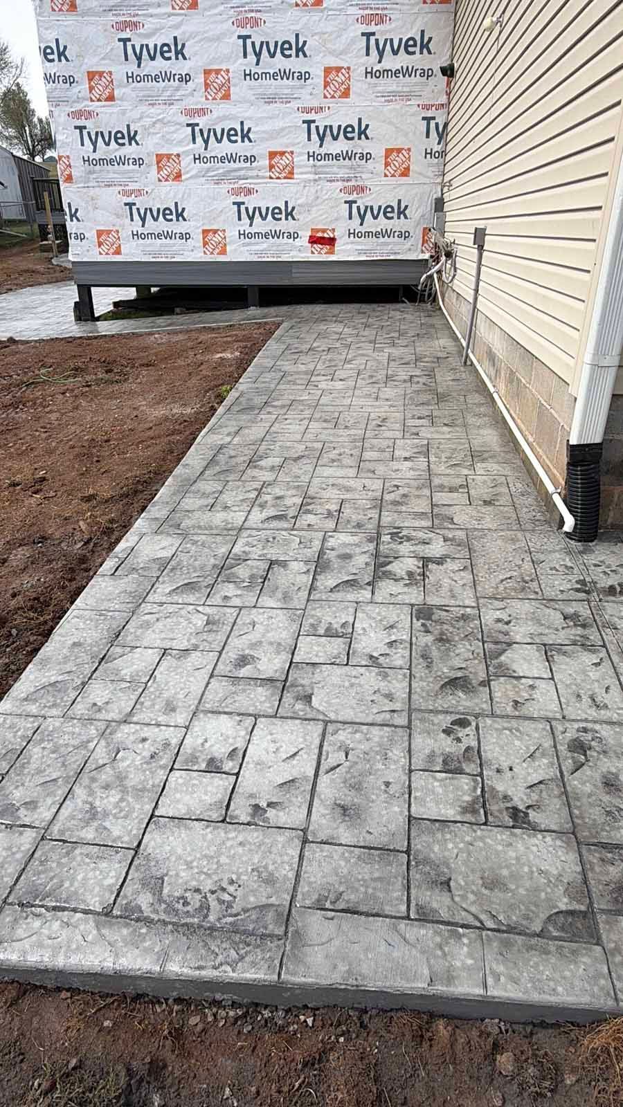Concrete walkway beside house under construction, bordered by soil and unfinished siding with Tyvek wrap in the background.