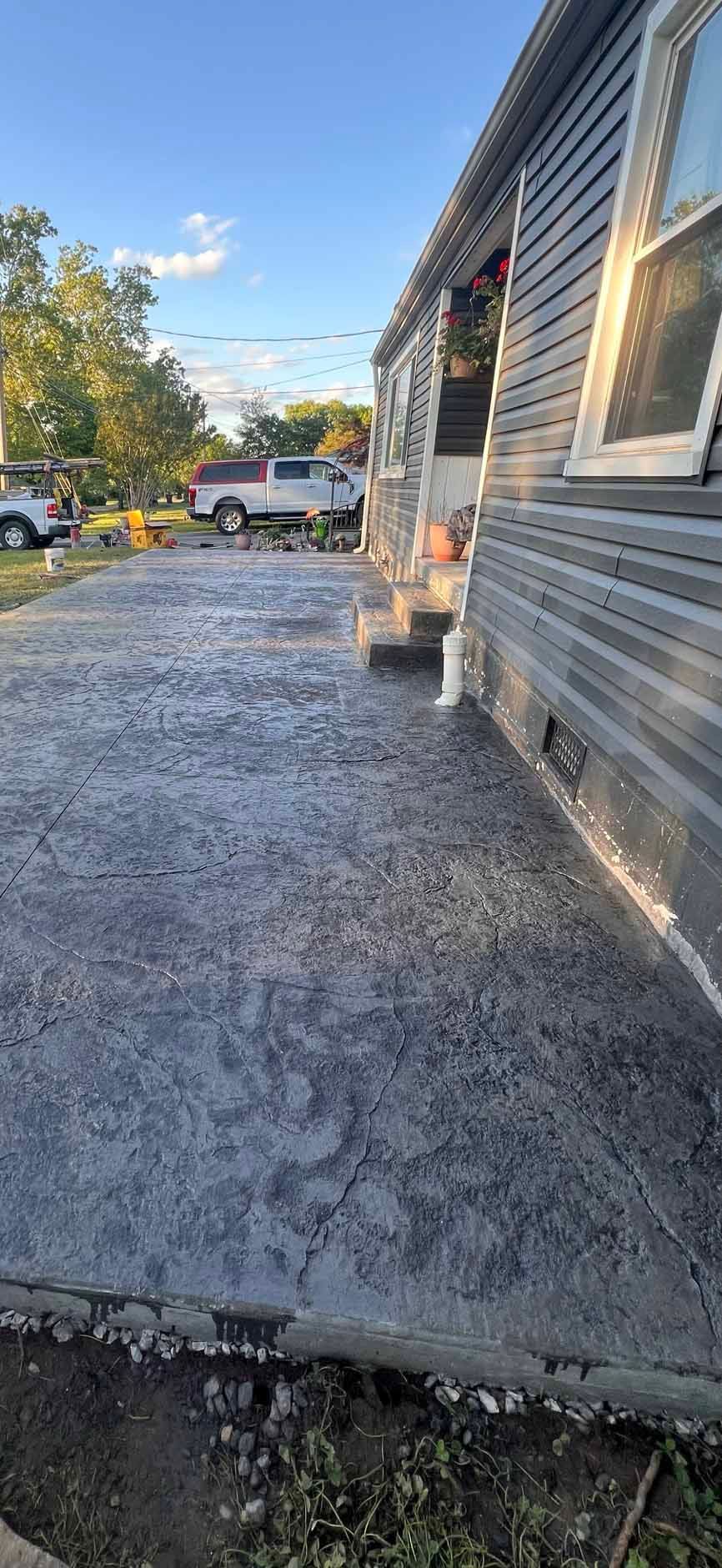 Gravel driveway beside a gray house with a porch, parked cars, and trees under a blue sky