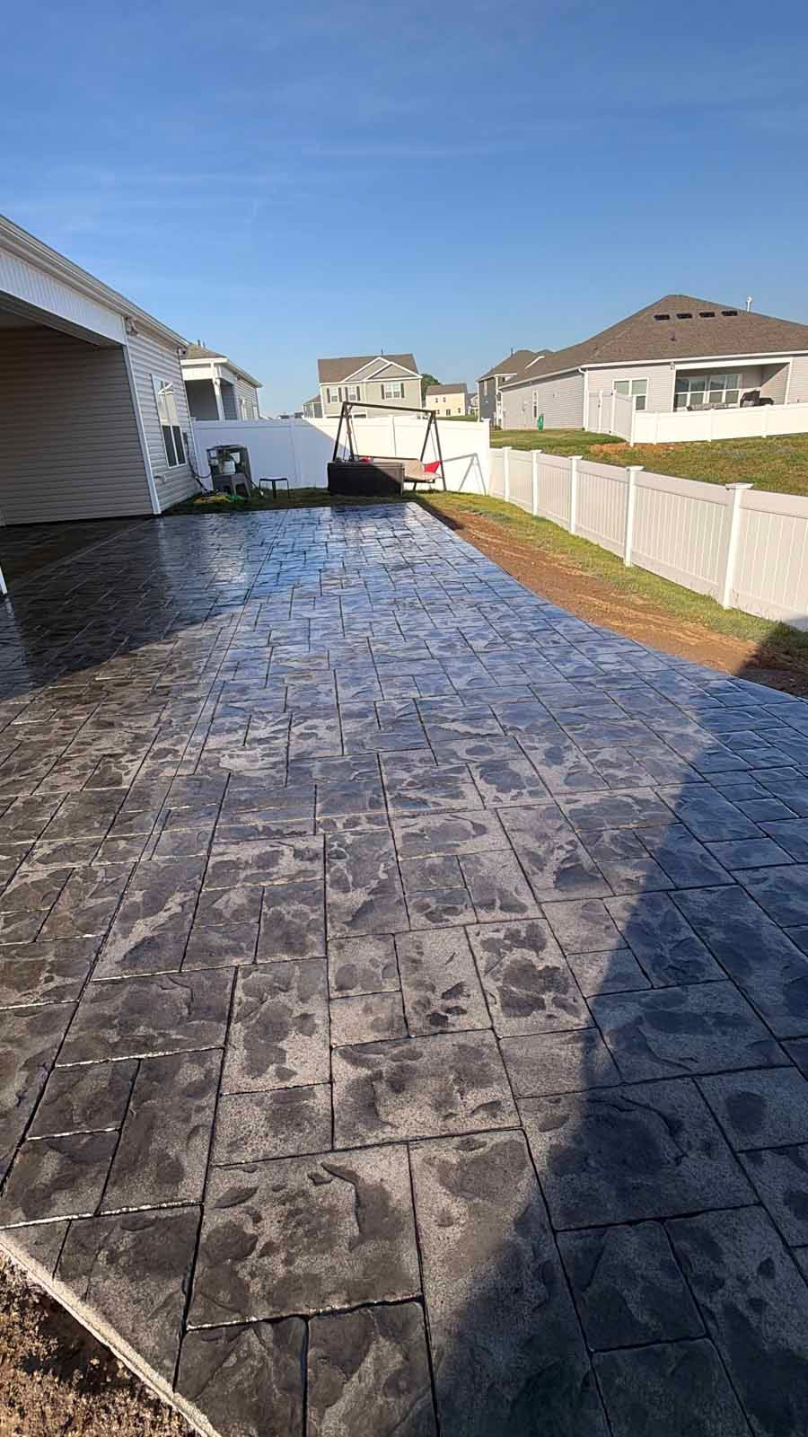Narrow backyard with dark patterned paving, bordered by white fences and houses under a blue sky
