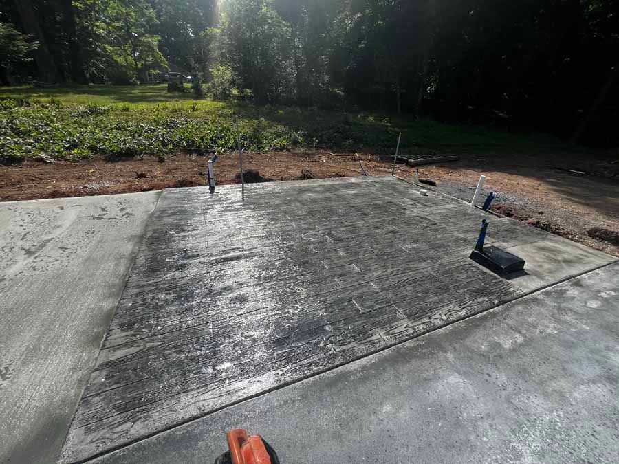 Concrete driveway edge with two small sprinkler heads beside a mulched garden bed and trees.