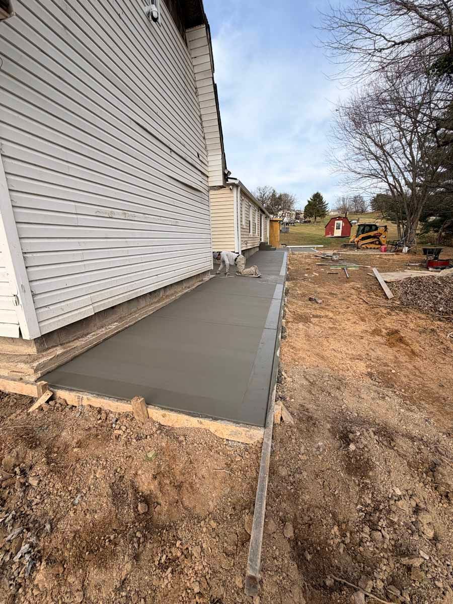 Fresh concrete walkway alongside a white house under construction, with dirt and a yard in the background