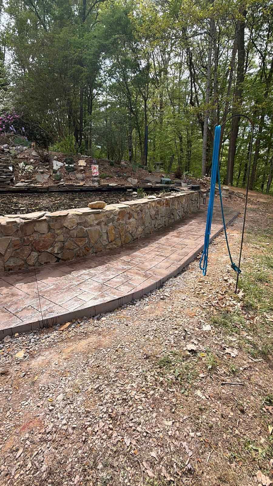 Curved stone retaining wall and path in a wooded outdoor area, with a blue post in the foreground