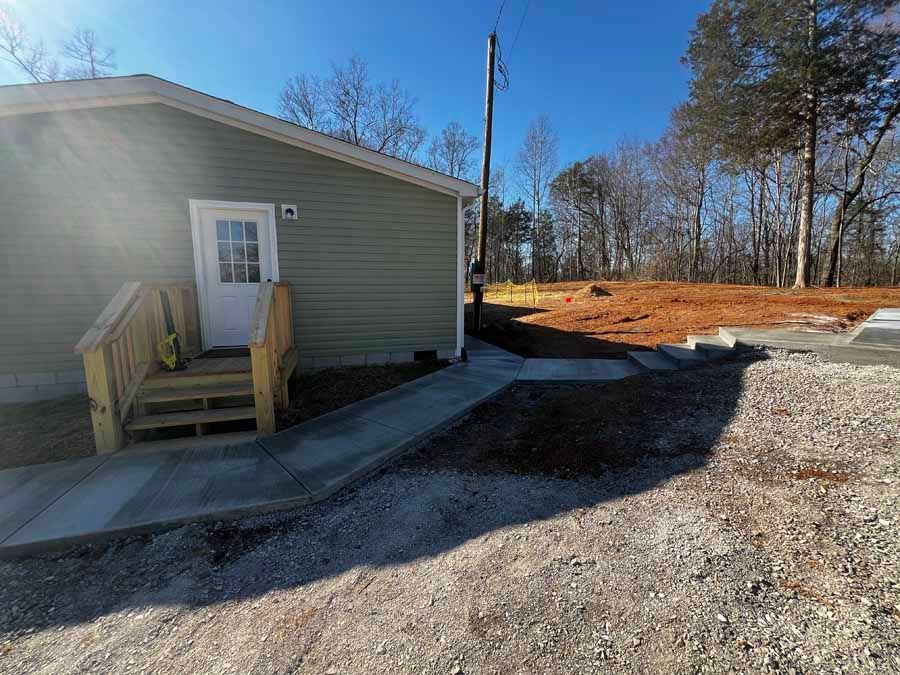 Small beige building with a wooden ramp and gravel driveway in a sunny, wooded area
